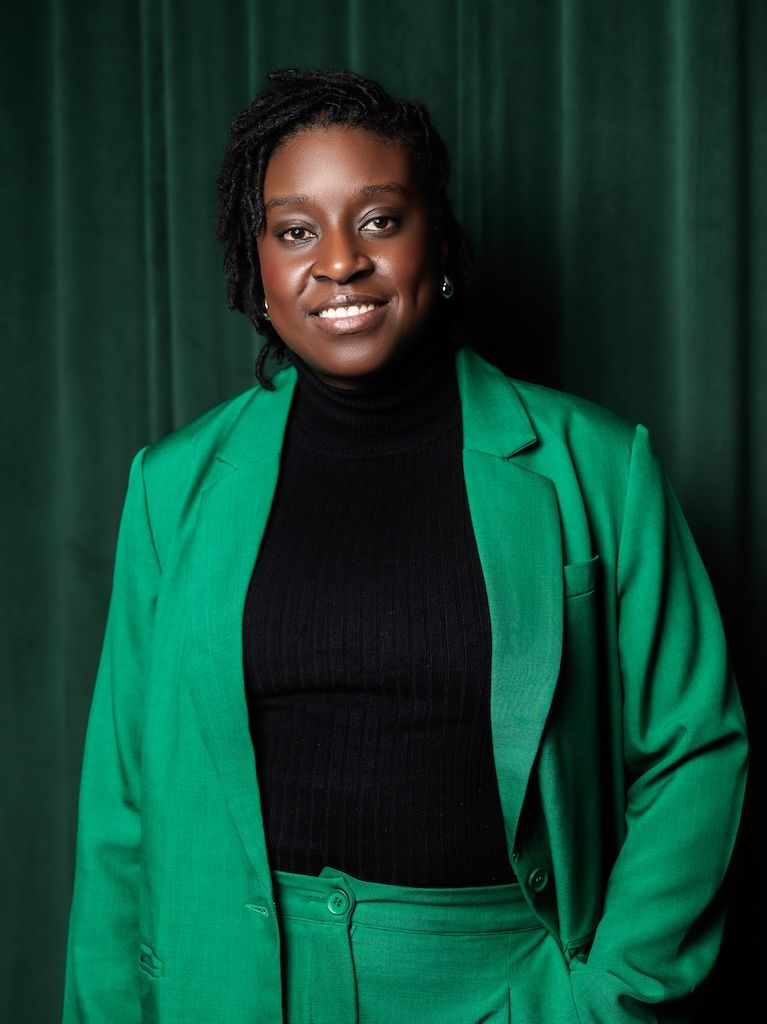 Smiling woman in a green suit and black turtleneck standing before a dark green curtain