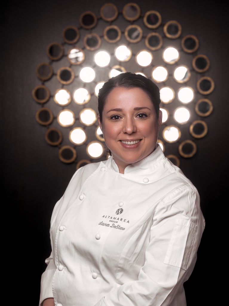 Smiling chef in white coat embroidered "Lauren DeStefano" before a glowing circular light fixture